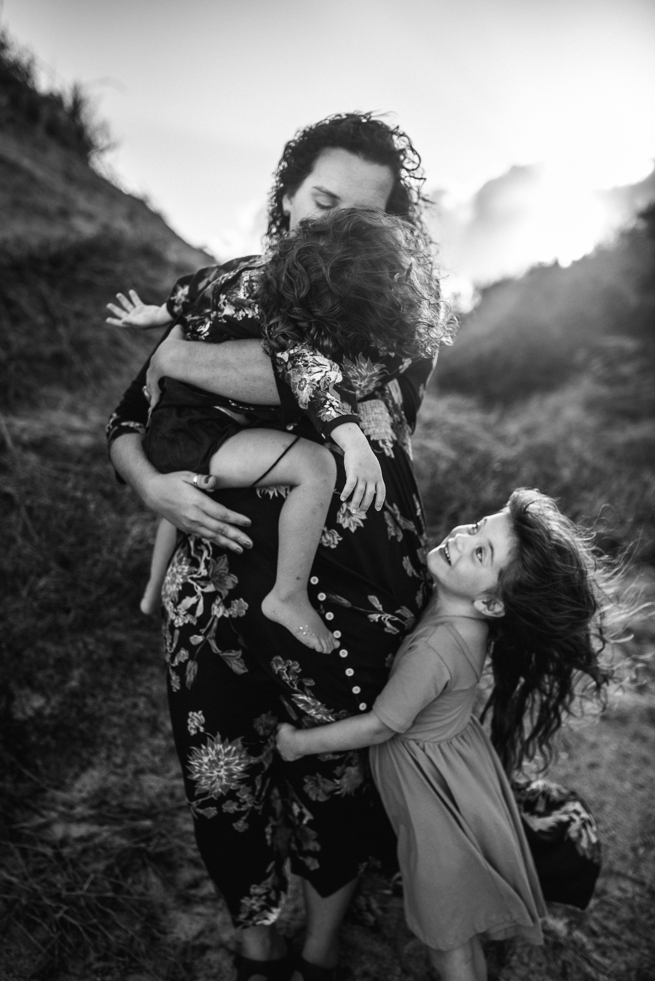 full of emotions family portrait of mom and two daughters with dunes in the background as sun goes down and wind tosses their hair as they hug tightly