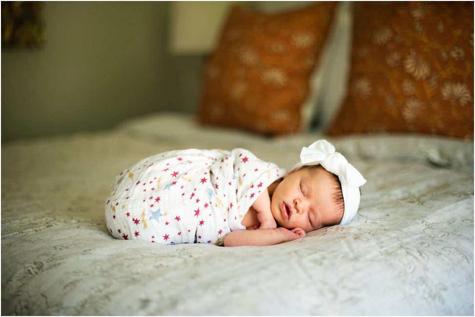 “Newborn baby girl wrapped in a star-patterned swaddle with white bow headband, sleeping peacefully on a bed with warm accent pillows in a cozy bedroom.”