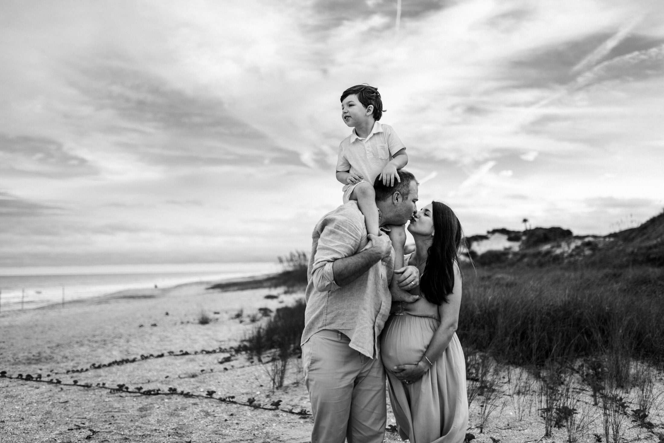 Family posing for photo with parents kissing while mom holds baby bump and dad holds small child on his shoulders in a family photoshoot in Jacksonville FL