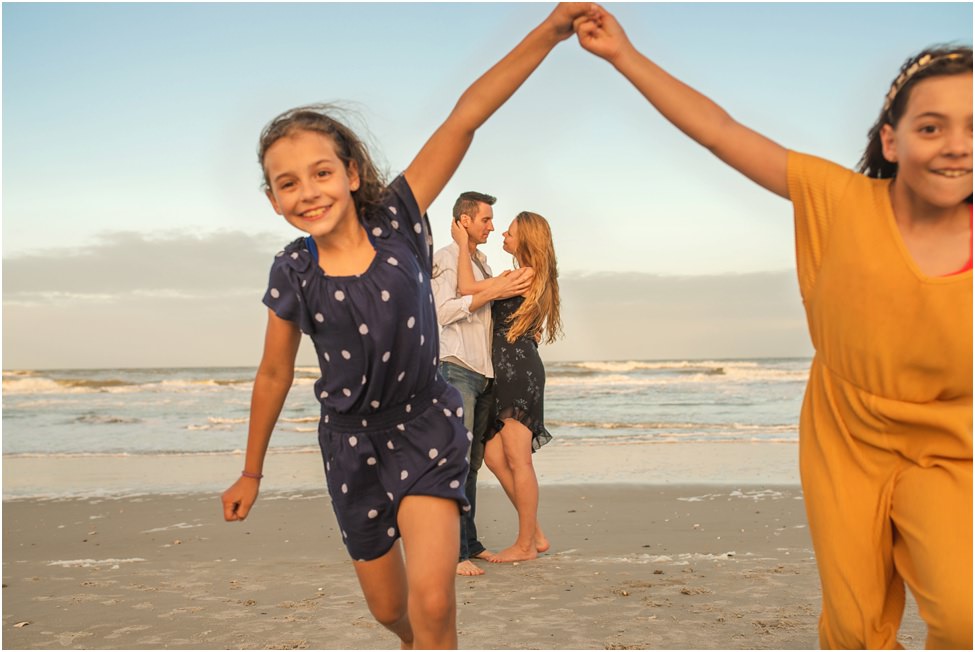 Children playing on the beach with parents hugging in the background in a family photoshoot in Jacksonville FL
