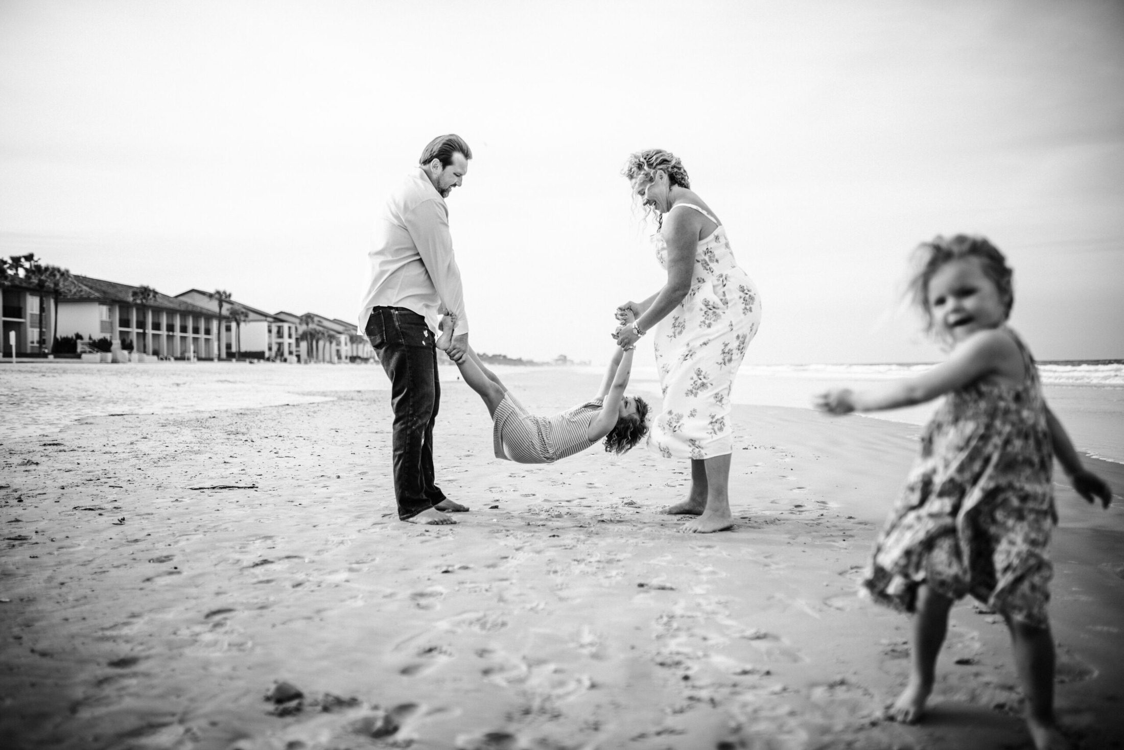 Parents playing with one child while another child runs in the background on the beach in a family photoshoot in Jacksonville FL