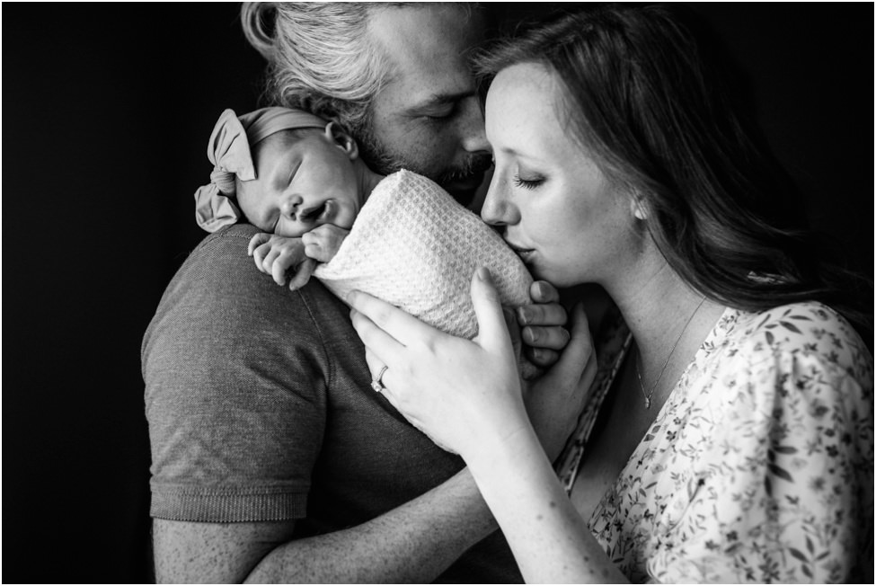 “Parents embracing their newborn baby girl wrapped in a blanket, mother kissing baby’s head while father holds her close, black and white lifestyle portrait capturing tender family connection.”