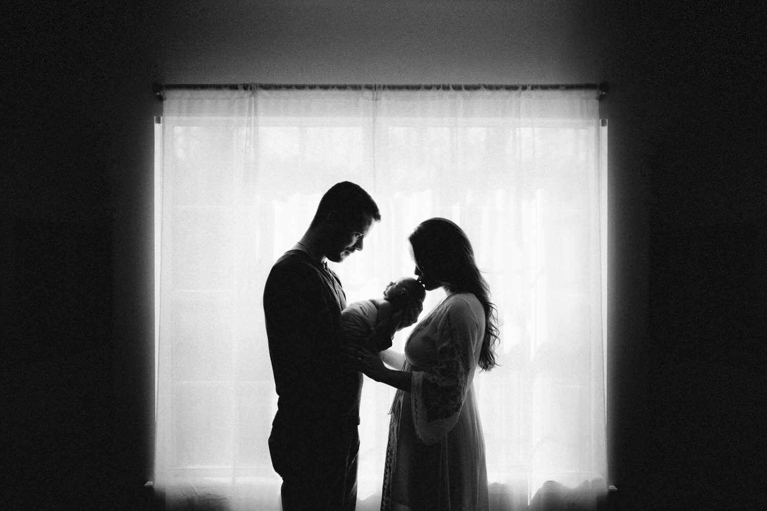 “Silhouette of parents holding newborn baby in front of sheer window curtains during intimate in-home session”