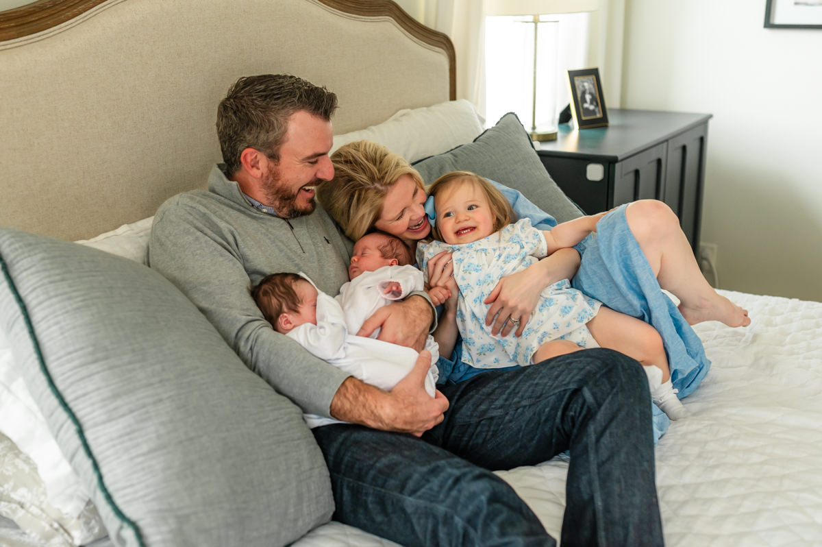 “Happy family of four with newborn twins and toddler snuggling on bed during lifestyle photo session in Jacksonville home”