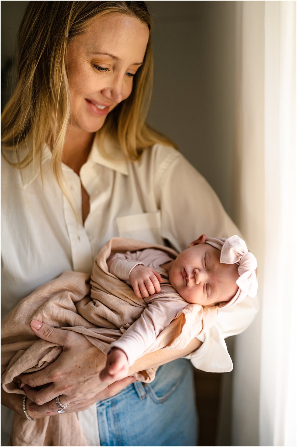 “Mother smiling down at newborn baby girl in pink wrap and bow during natural light lifestyle session in Jacksonville home”