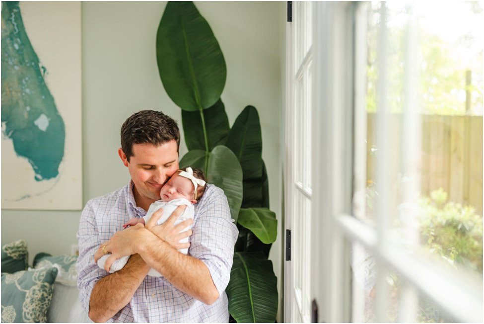 “Father holding newborn baby girl with bow headband close to his cheek during in-home newborn session in Jacksonville, Florida”