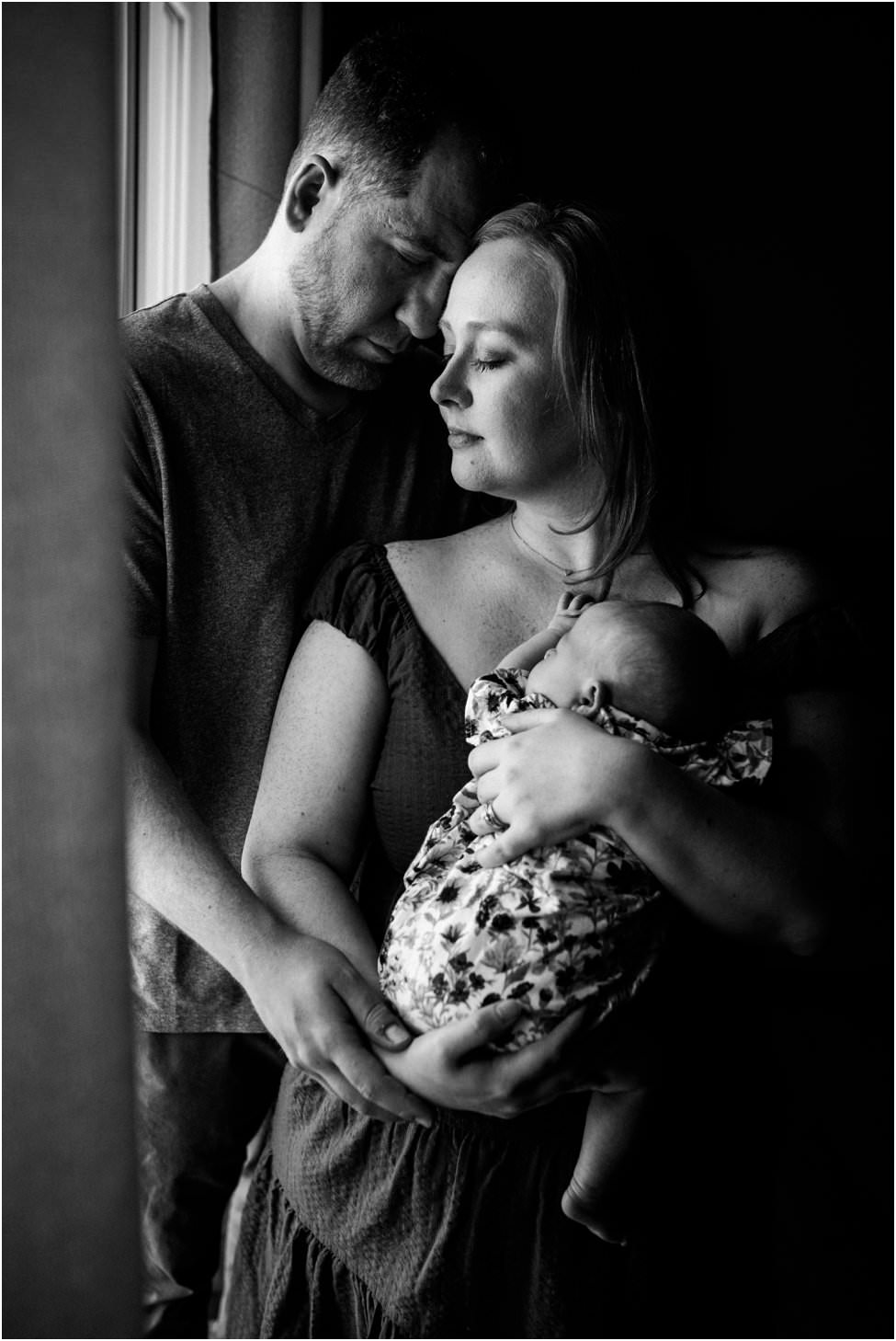 “Mother and father standing by a window, gently holding their newborn baby close during an intimate lifestyle session in Jacksonville by Monika Colichio Photography”