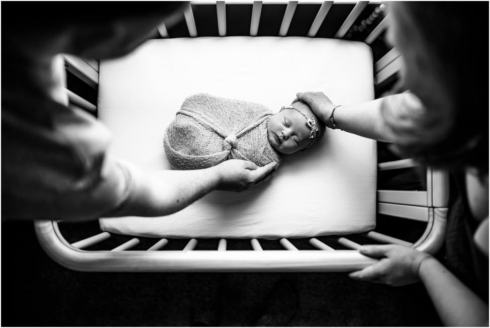 “Parents placing their swaddled newborn baby with a delicate headband into a crib during an in-home lifestyle session in Jacksonville by Monika Colichio Photography”