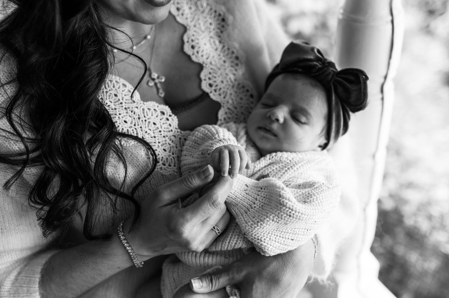 “Mother holding her sleeping newborn dressed in a cozy sweater and bow headband, gently touching hands during an in-home lifestyle session in Jacksonville by Monika Colichio Photography”
