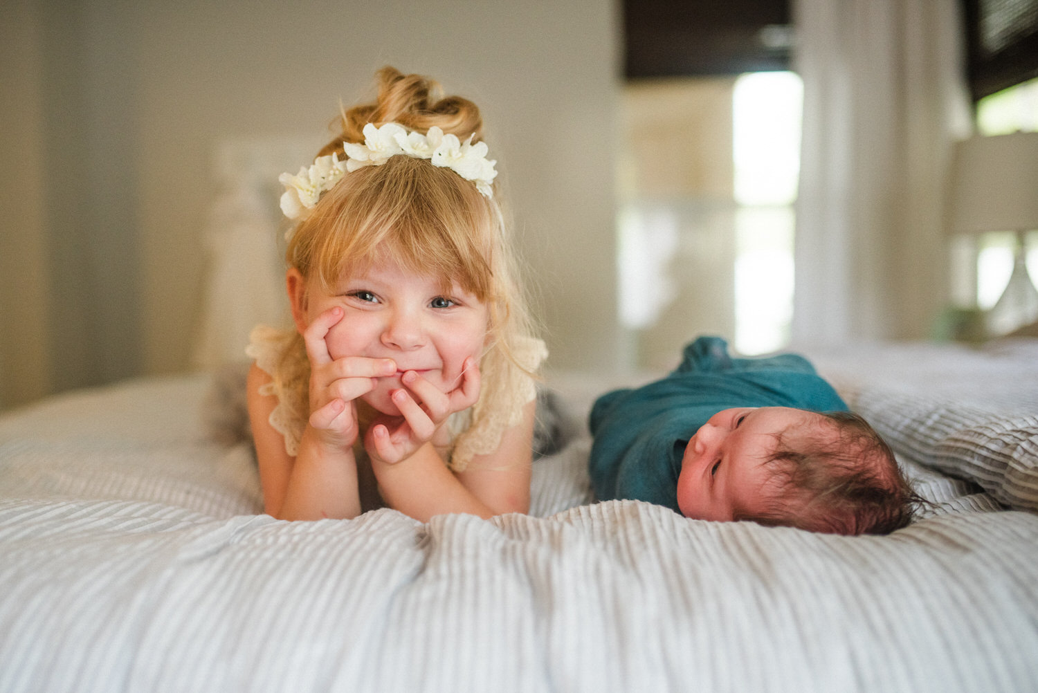 “Big sister smiling playfully next to her swaddled newborn sibling on a bed during an in-home lifestyle session in Jacksonville by Monika Colichio Photography”