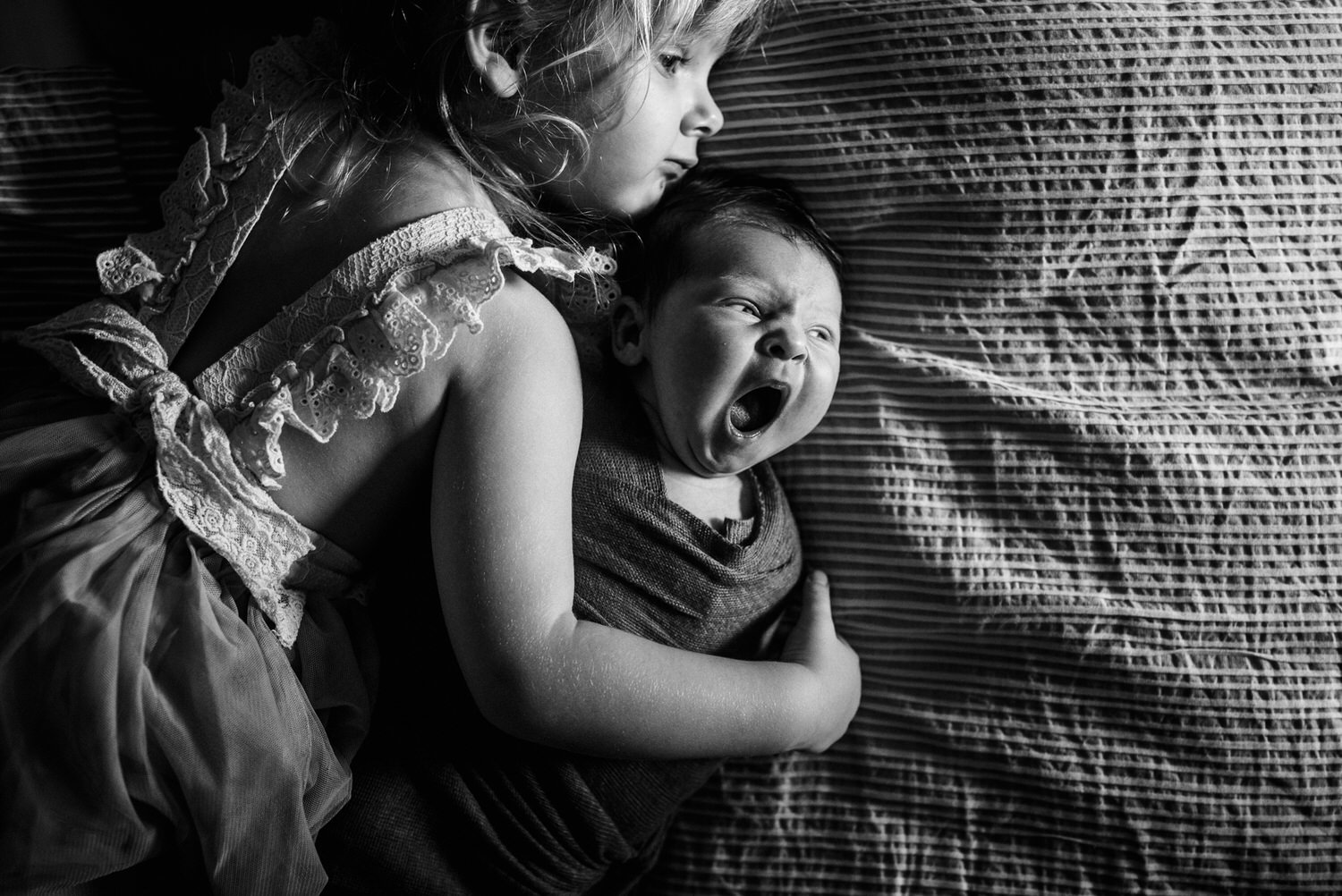 “Big sister cuddling her yawning newborn sibling on a bed during an intimate lifestyle session in Jacksonville by Monika Colichio Photography”
