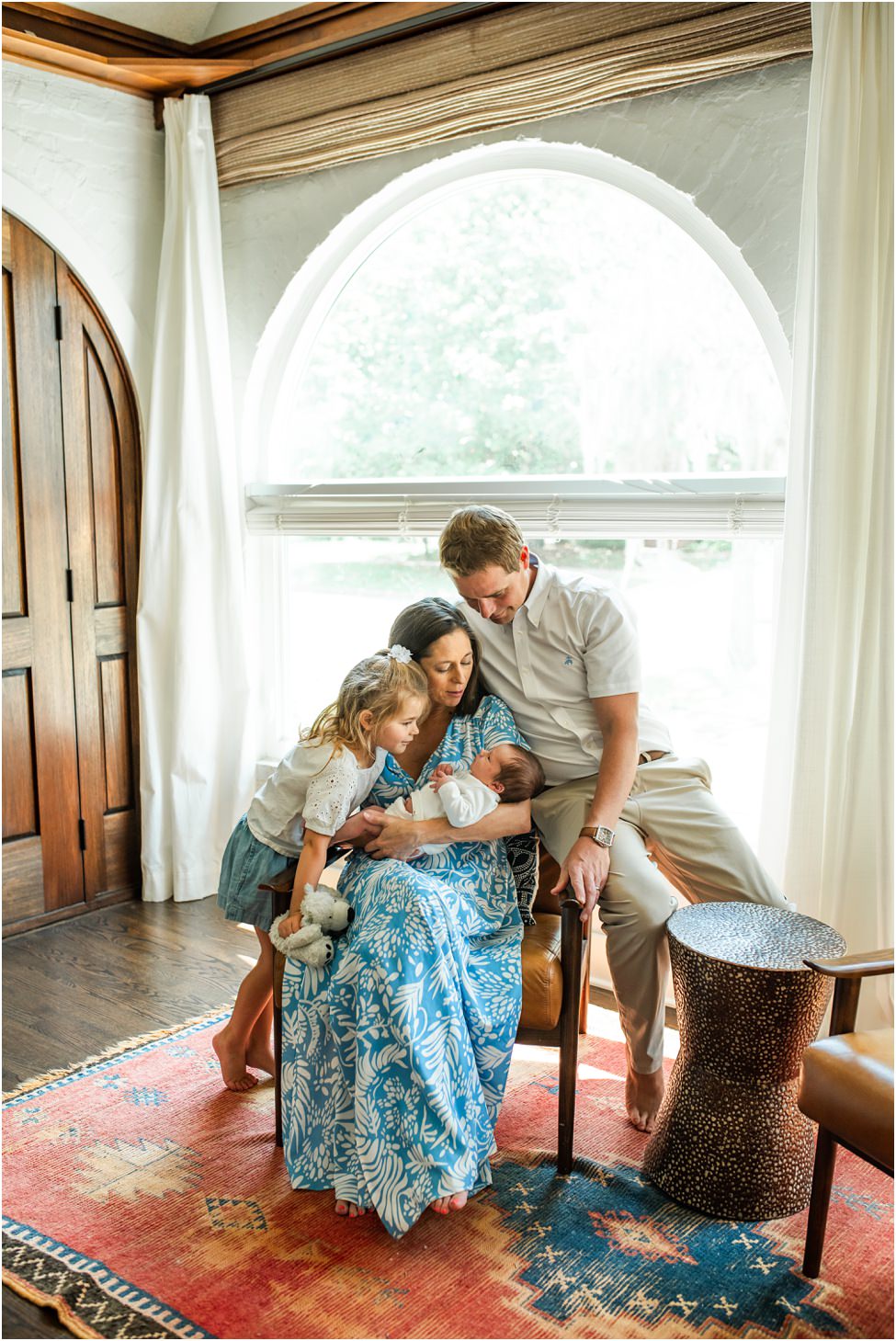 “Family gathered by a large arched window, with mother holding newborn, father leaning close, and big sister looking on during an in-home lifestyle session in Jacksonville by Monika Colichio Photography”