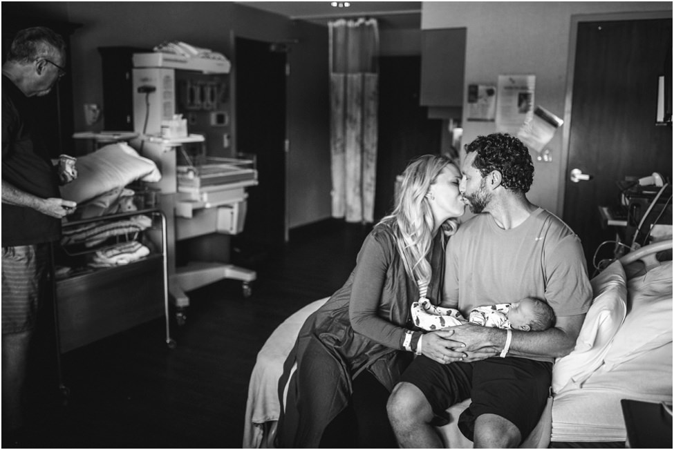 “Emotional mother kissing new dad holding a newborn son while sitting in the hospital bed during first 48 hours Jacksonville hospital newborn session by Monika Colichio Photography