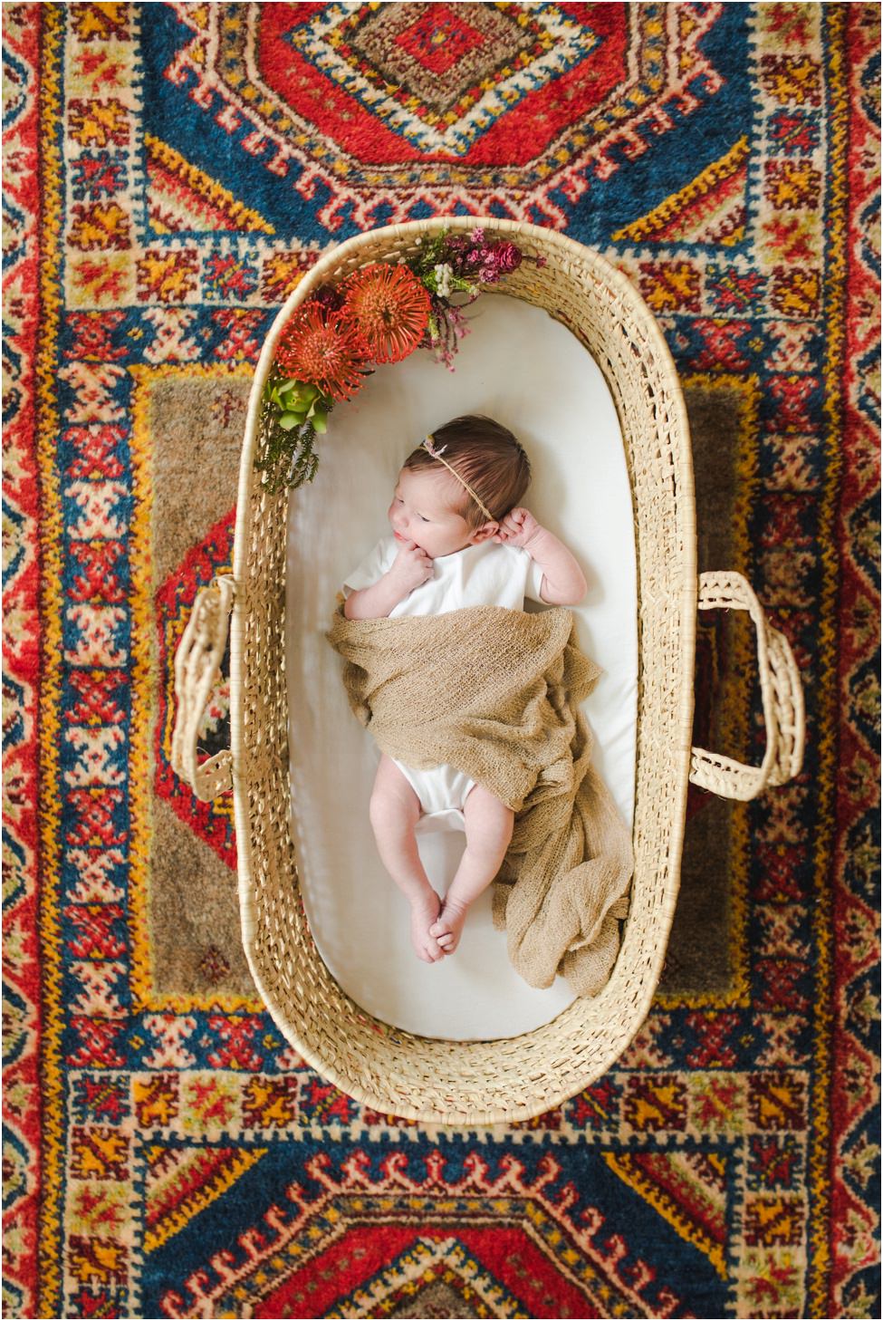 “Newborn baby in a woven basket with flowers, wrapped in a light brown blanket, lying on a colorful patterned rug during a lifestyle session in Jacksonville by Monika Colichio Photography”