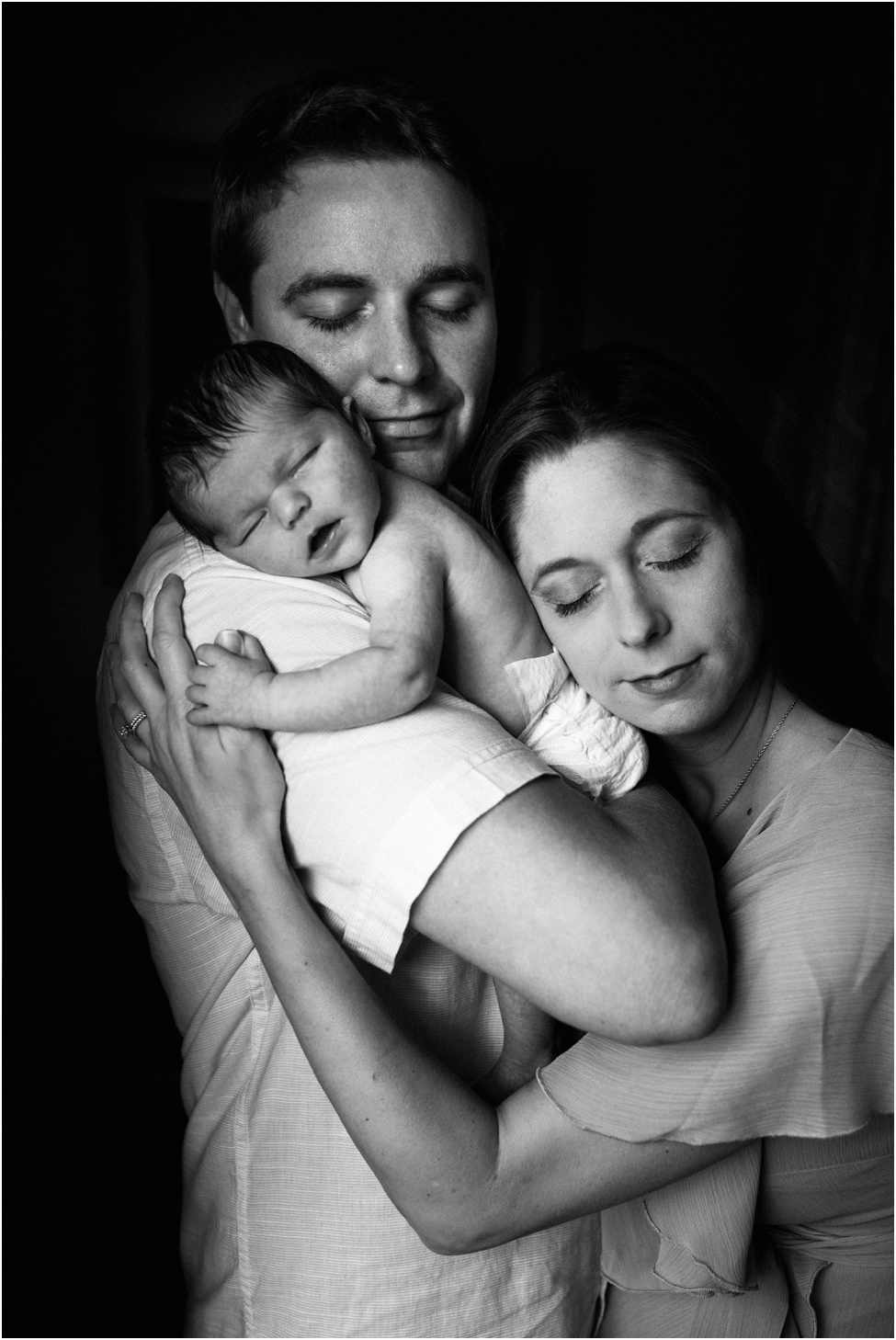 “Parents embracing each other with their newborn baby resting peacefully on father’s shoulder during an intimate lifestyle session in Jacksonville by Monika Colichio Photography”