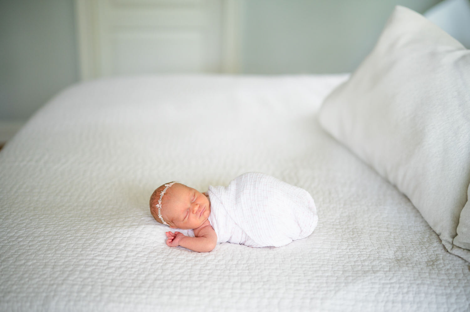“Newborn baby girl swaddled in white with a delicate headband sleeping on a bed during a light-filled lifestyle session in Jacksonville by Monika Colichio Photography”