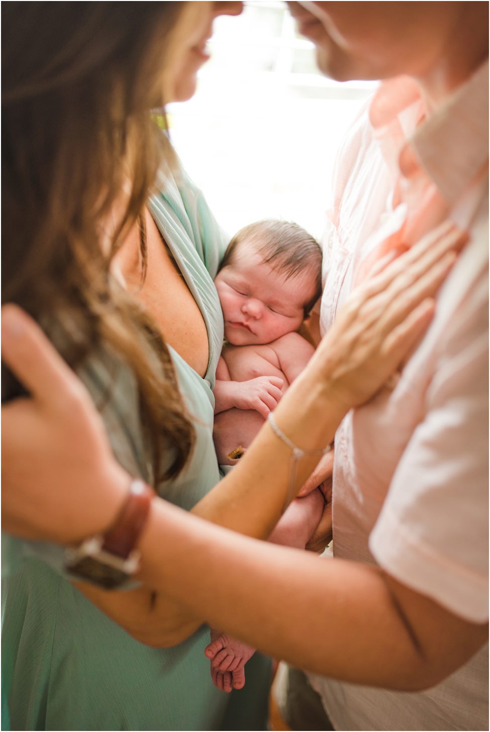 “Newborn baby with full dark hair sleeping peacefully in mother’s arms, with parents embracing closely and touching shoulders, captured in a warm lifestyle family portrait.”