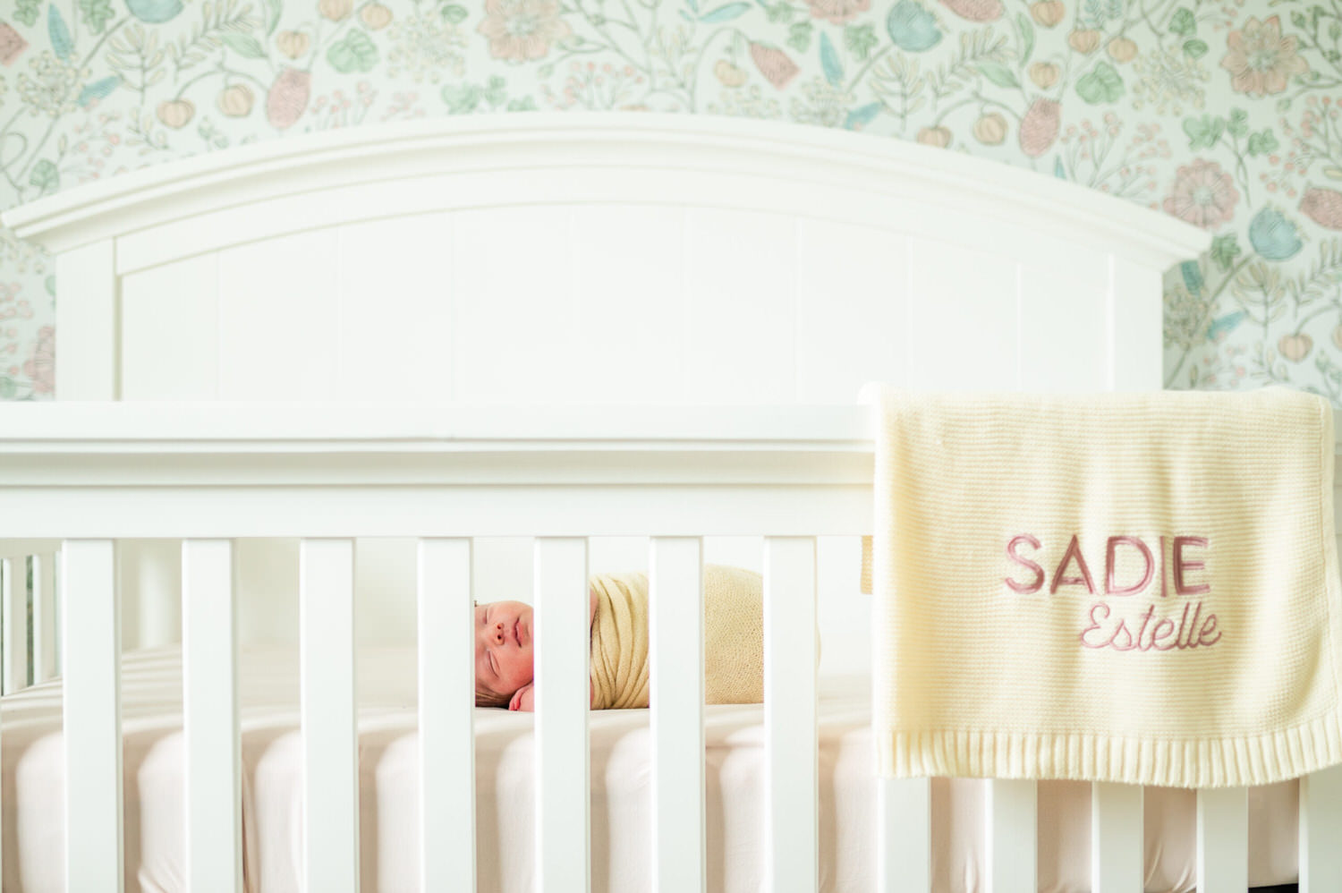 “Newborn baby wrapped in a yellow swaddle sleeping in white crib with floral wallpaper and personalized blanket during a lifestyle session in Jacksonville by Monika Colichio Photography”