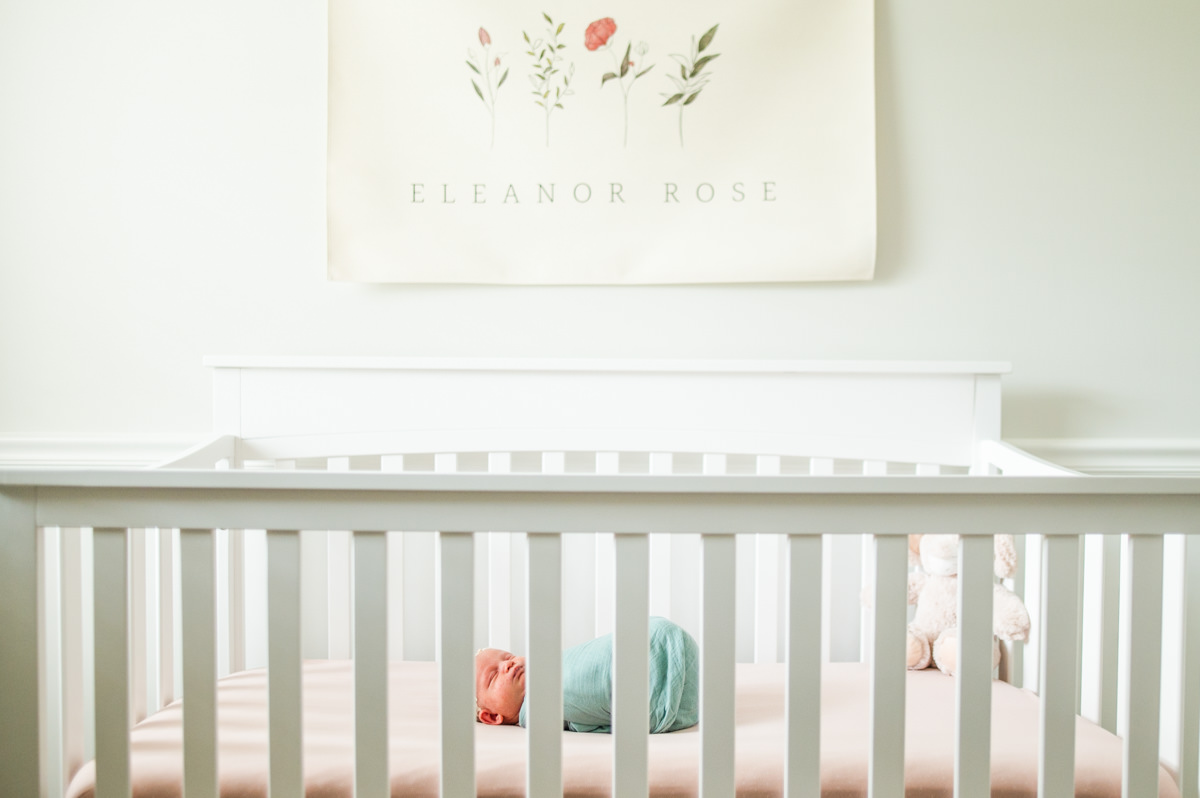 Newborn baby swaddled in a soft green wrap lying in a white crib beneath a floral name sign during a lifestyle newborn session in Jacksonville by Monika Colichio Photography