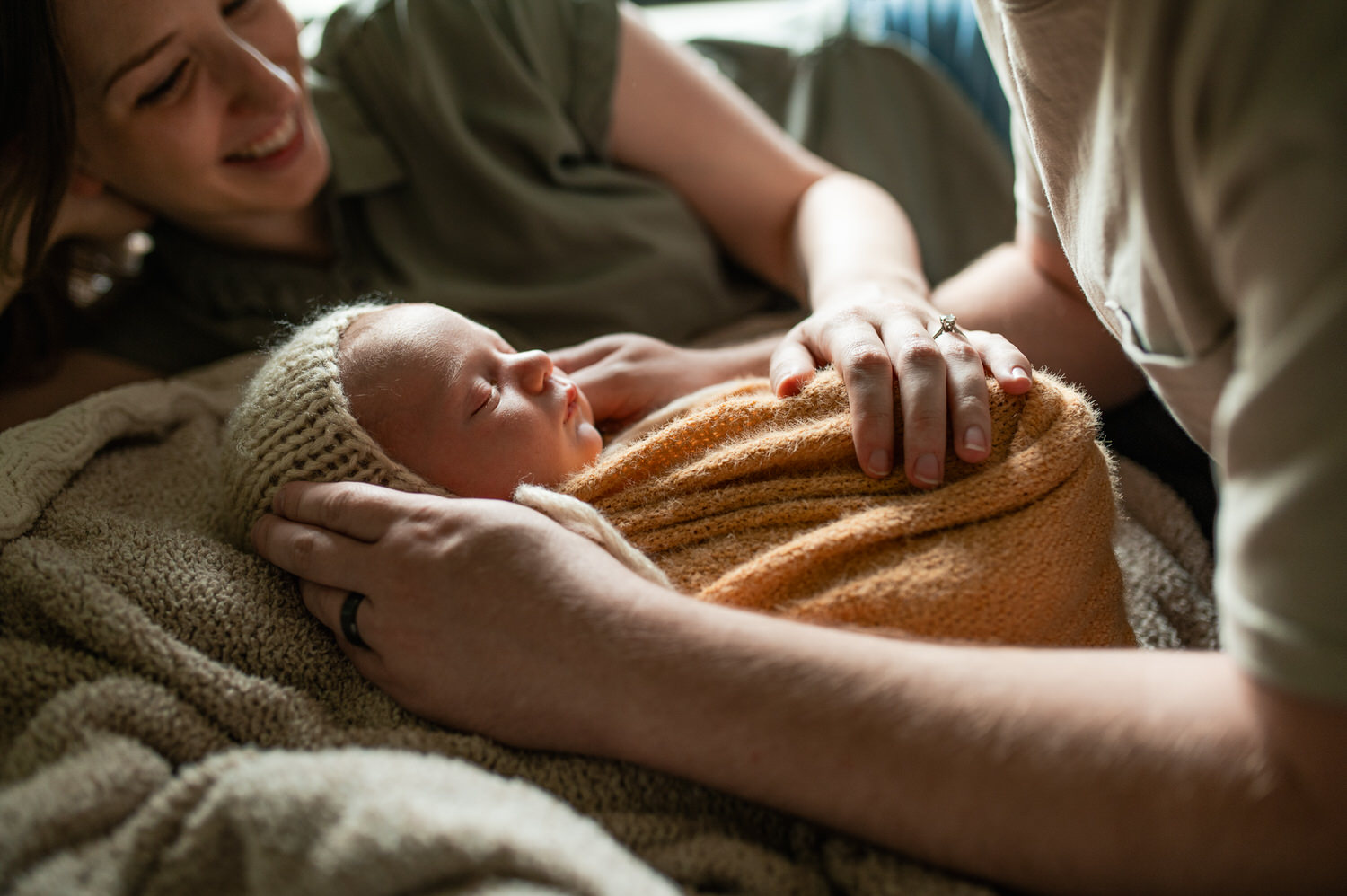 “Newborn baby wrapped in a soft orange blanket and knit bonnet, held lovingly by parents during a cozy lifestyle session in Jacksonville by Monika Colichio Photography”
