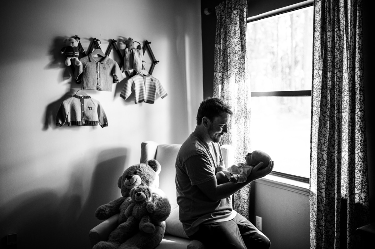 “Father smiling down at his newborn baby by the nursery window during an intimate lifestyle session in Jacksonville, captured by Monika Colichio Photography”