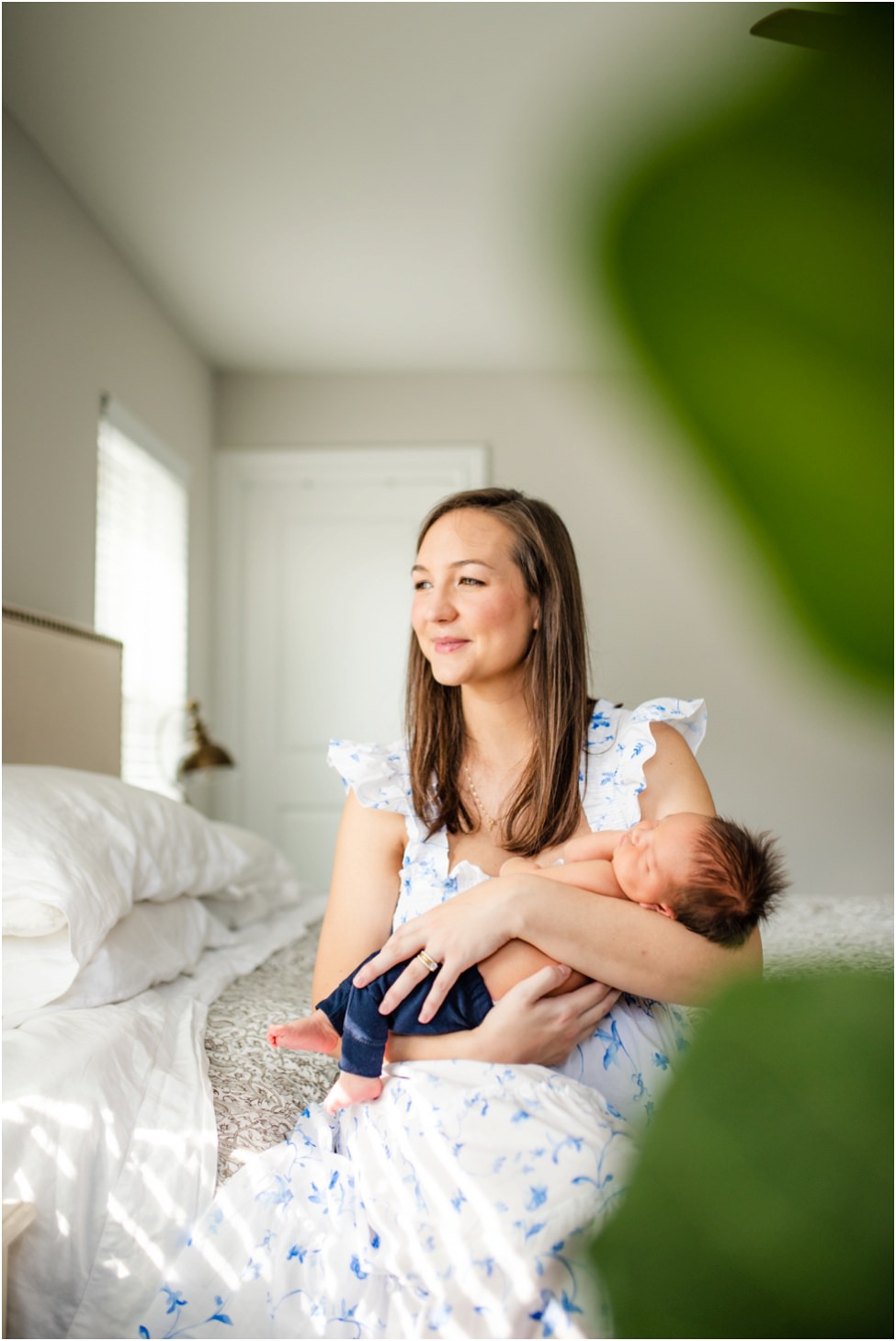 “Mother in a blue floral dress cradling her newborn baby on the bed during a bright, natural light lifestyle session in Jacksonville by Monika Colichio Photography”