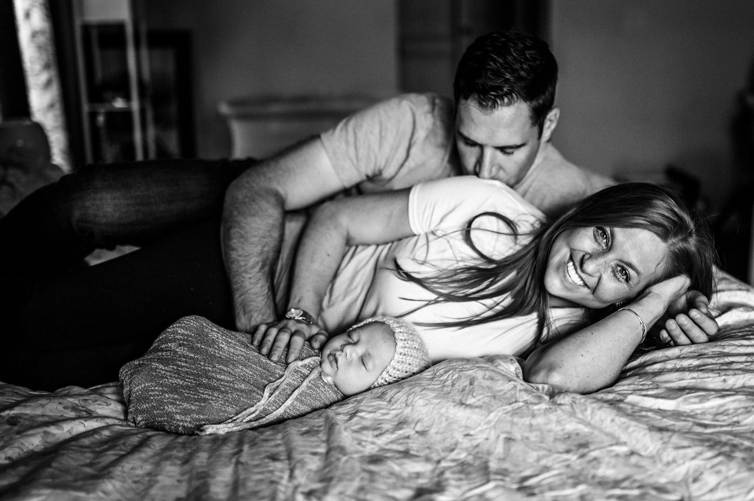 “Happy parents lying on a bed with their newborn baby wrapped in a knit swaddle and bonnet, mother smiling at the camera while father kisses her shoulder, captured in a cozy black and white lifestyle photo.”