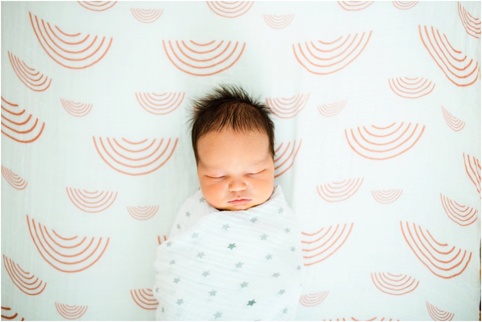 Newborn baby peacefully swaddled in a white blanket with gray stars, sleeping on a playful patterned sheet during an in-home lifestyle session in Florida by Monika Colichio Photography