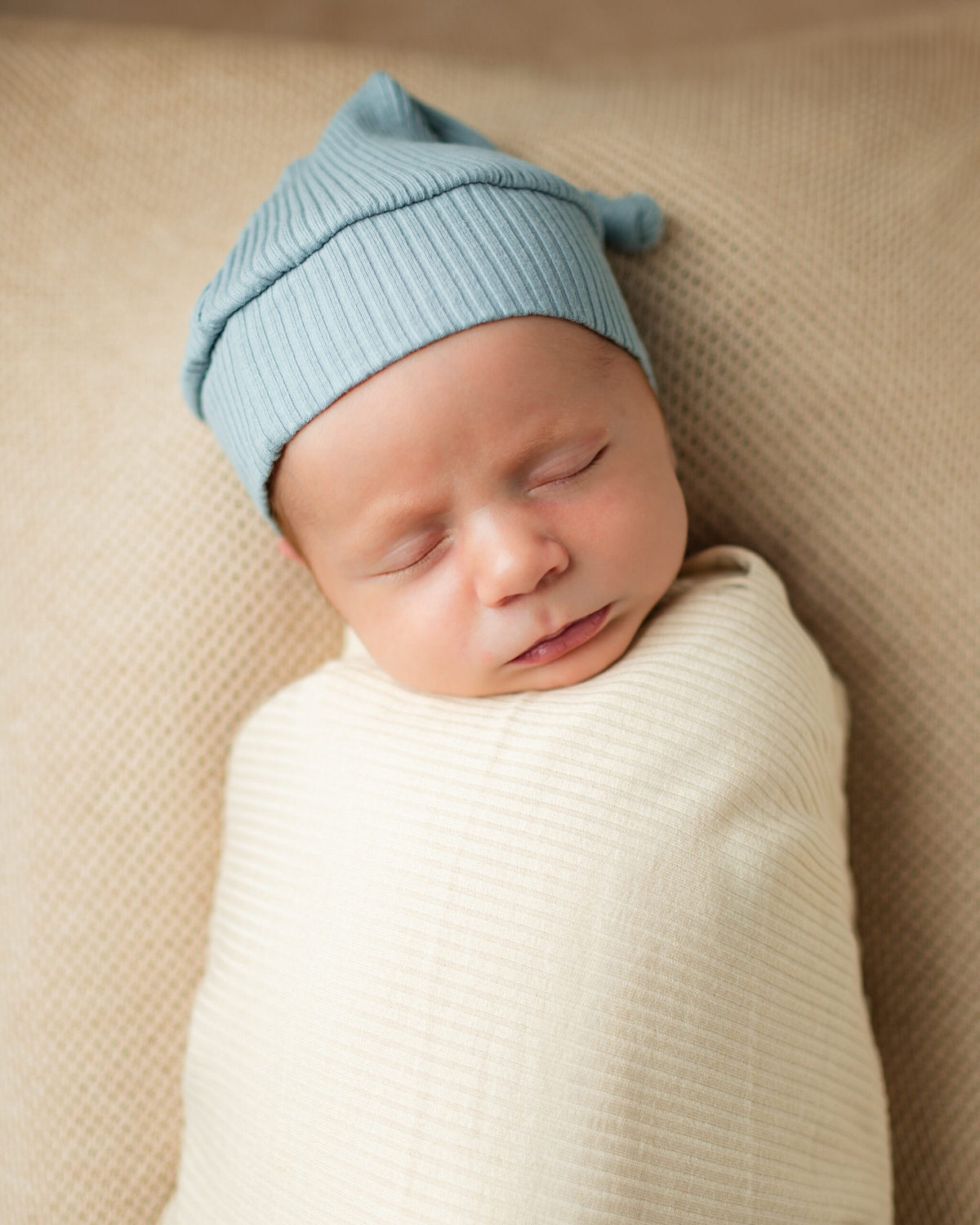 “Newborn baby peacefully sleeping, wrapped snugly in a cream-colored swaddle with a ribbed texture, wearing a light blue knit hat, resting on a beige textured blanket.”