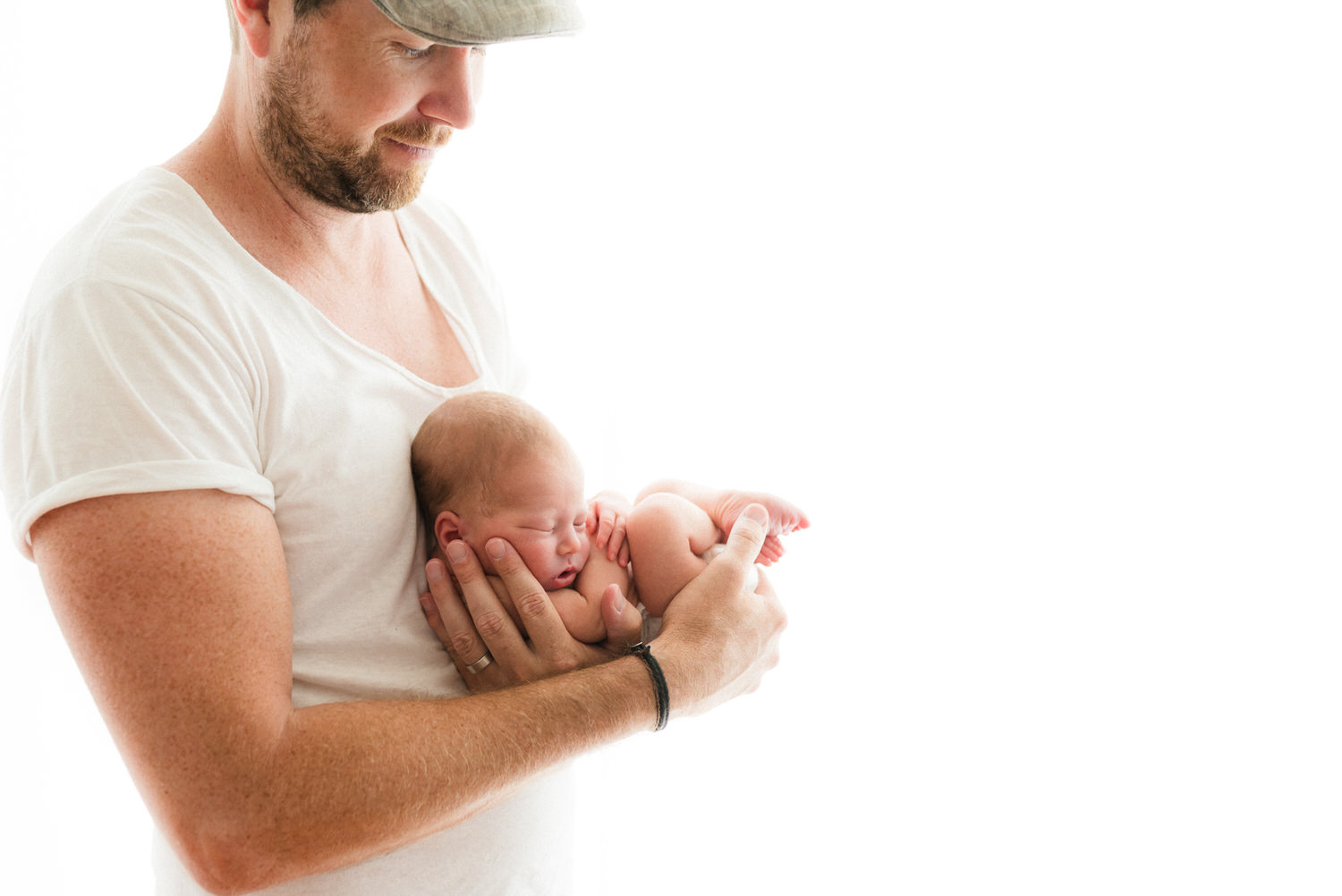 “Father wearing a white t-shirt and cap cradling his newborn baby against his chest, both softly lit against a bright white background.”