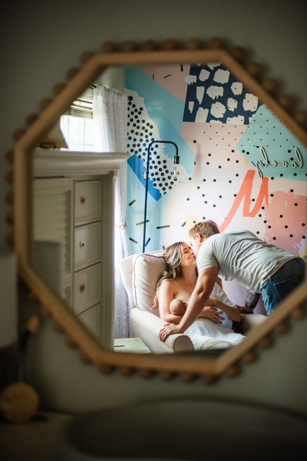 “Reflection in a decorative mirror of a mother breastfeeding her baby in a chair, as her partner leans in to kiss her, with a colorful abstract mural in the background.”