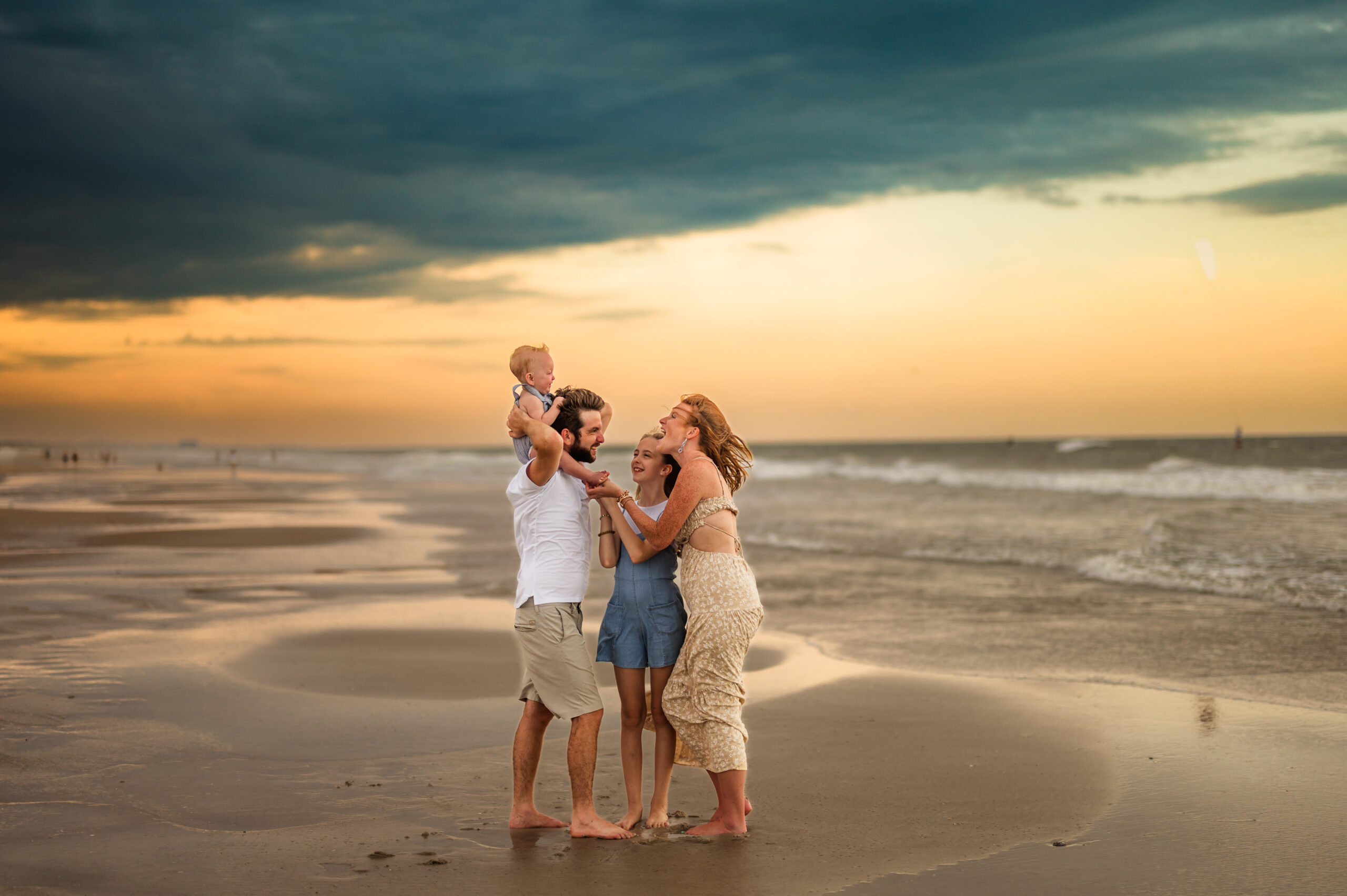 family picture of four with backdrop of beautiful stormy sky as golden sunset peaks from behind and wind tosses their hair and clothes by Atlantic Beach family photographer