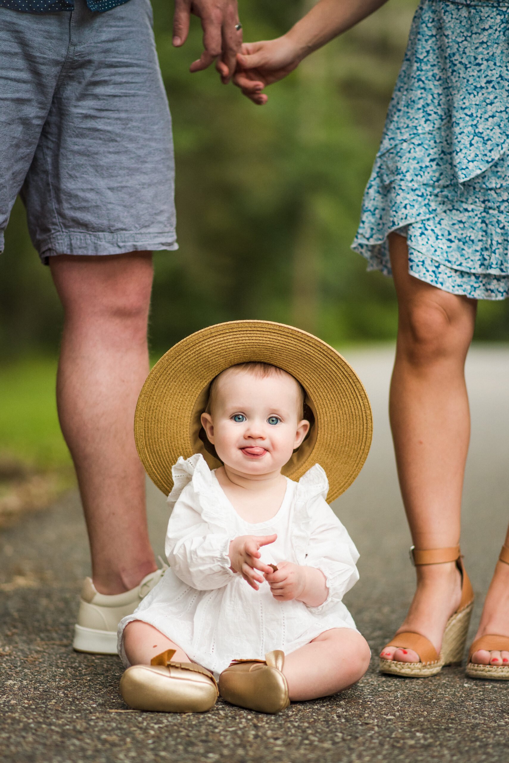 Adorable 1 year old in a summer hat sticking her tongue while sitting on the ground between her parents during family session by Jacksonville family photographer Monika Colichio