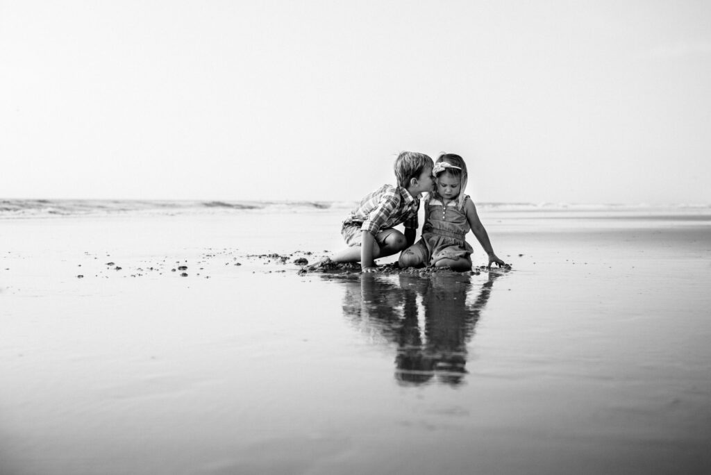 Two young children playing on the shoreline at low tide, with a tender moment captured by a Neptune Beach photographer in black and white