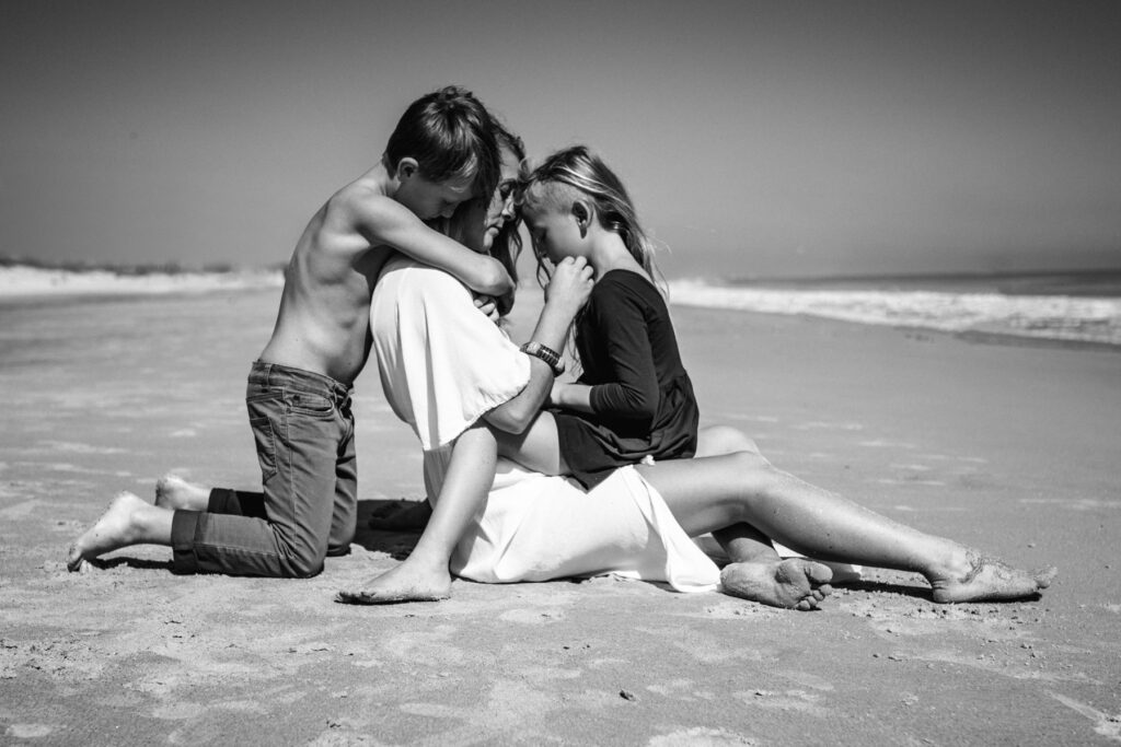 Emotive black and white photo of mom and kids hugging in the full sun as they sit on the beach by Ponte Vedra photographer